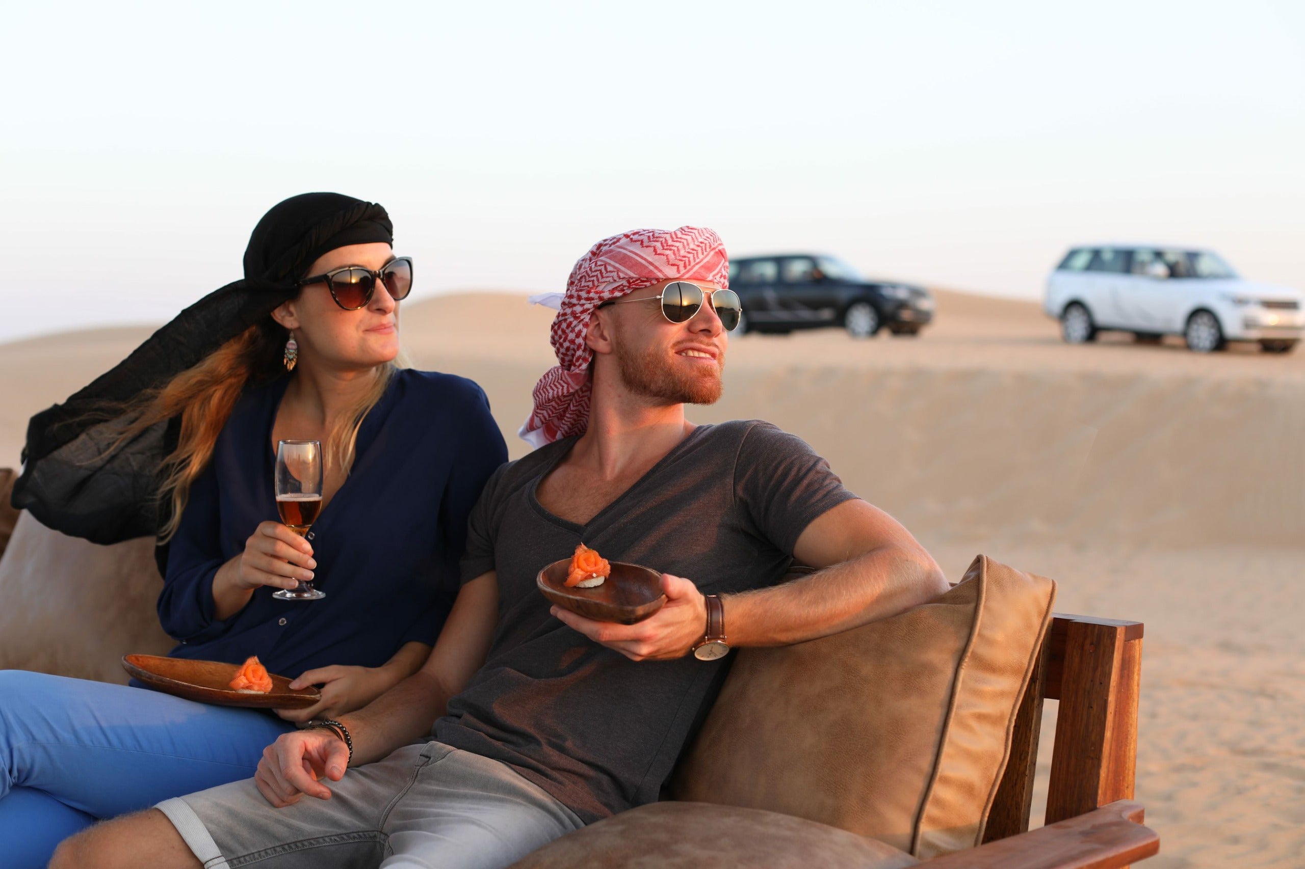 Two people sitting on a couch in the desert with vehicles in the background at platinum desert safari Dubai.