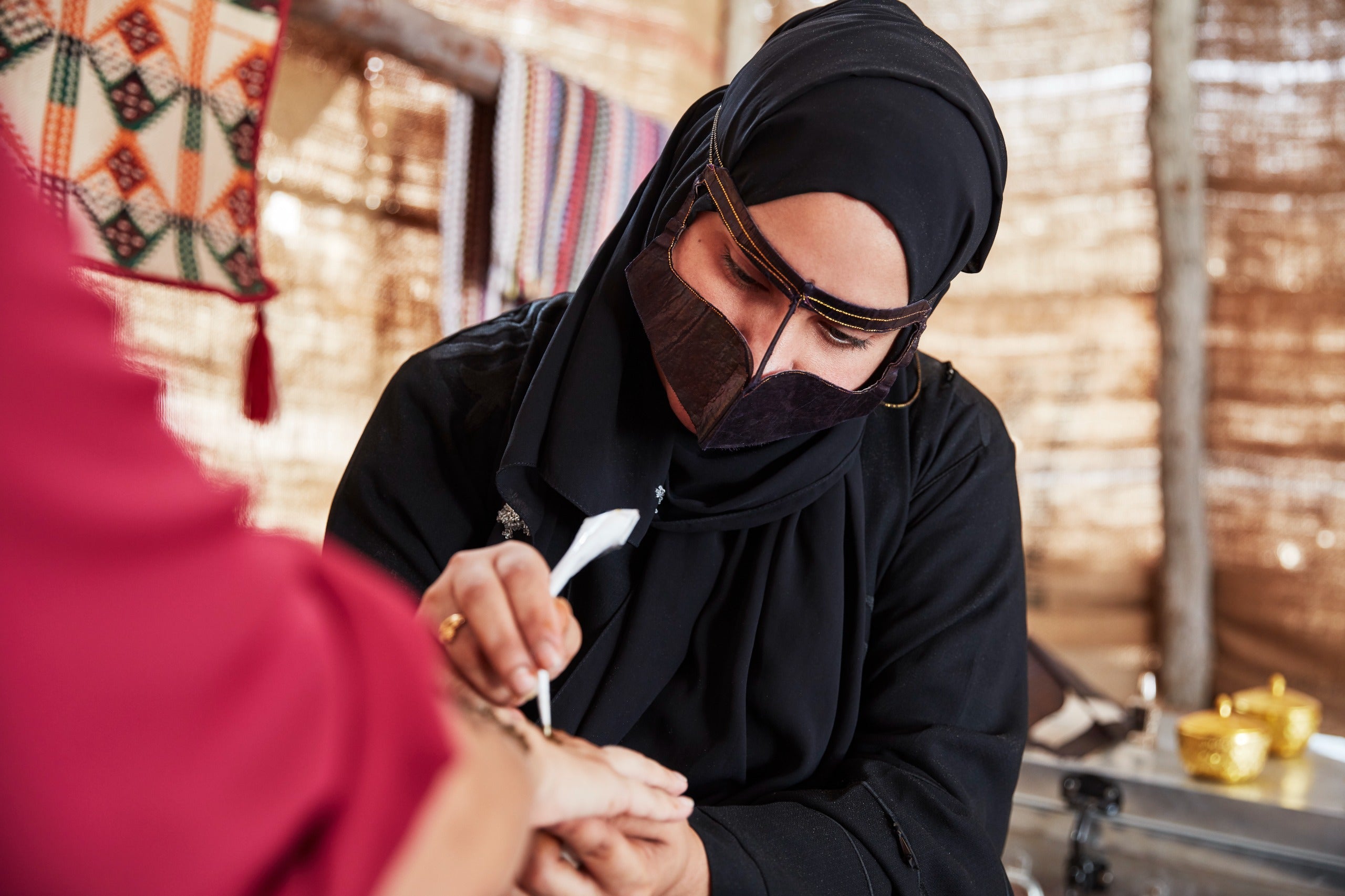 Woman in black hijab and face mask applying makeup to another person's face in a traditional setting.