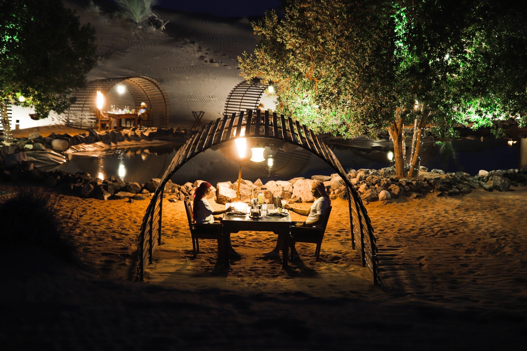 Dining table set up under a wooden canopy on a sandy beach at night.