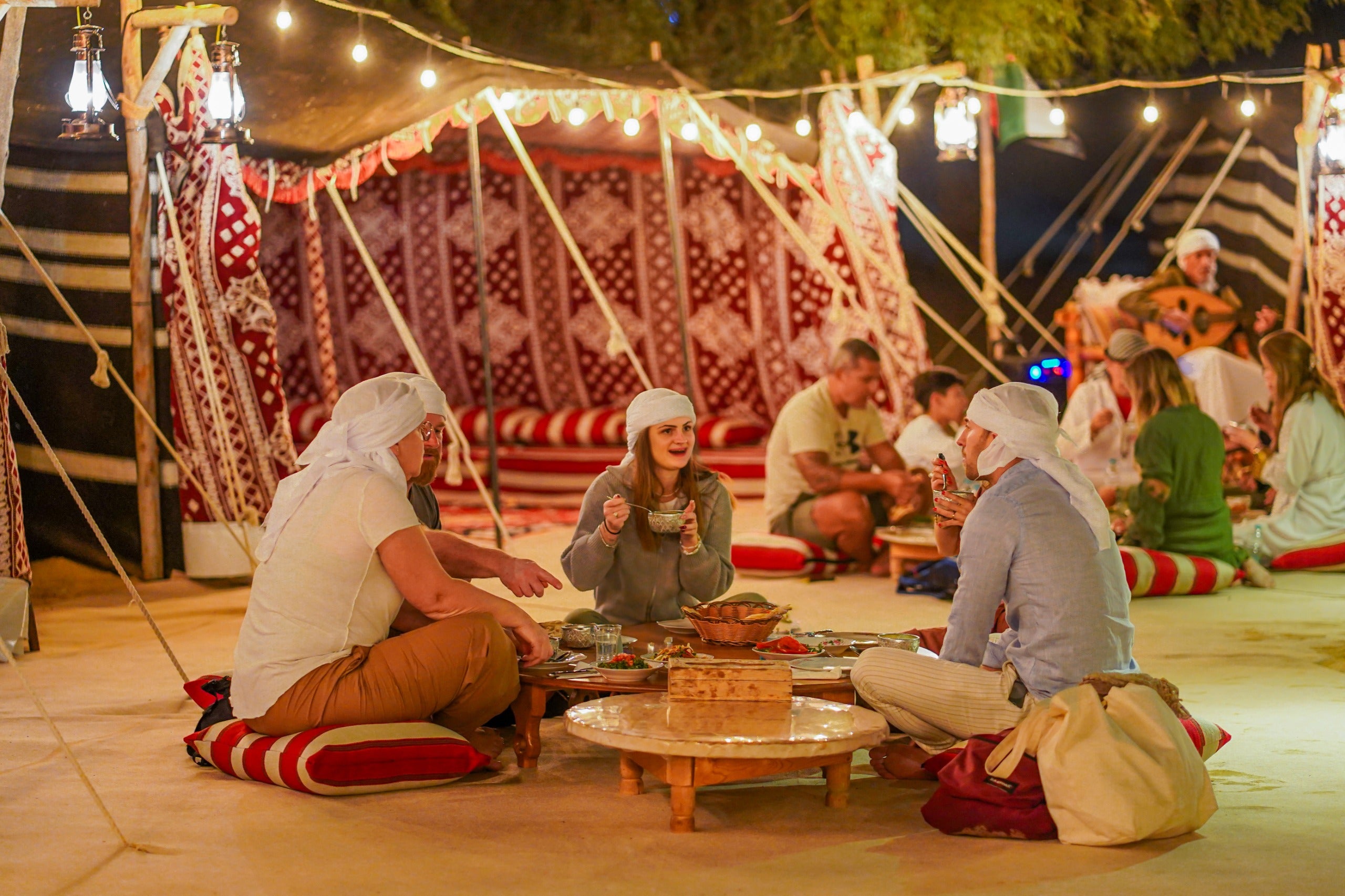 Group of people sitting on the ground around a small table with food, in a tented outdoor setting.