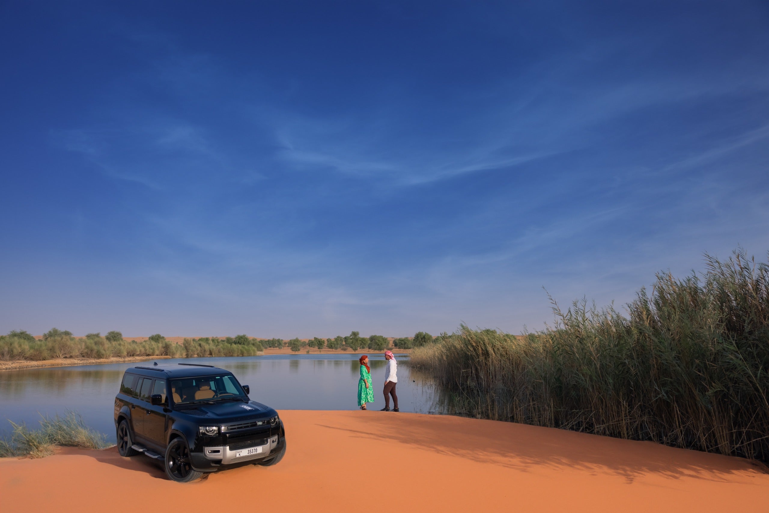 Two people standing near a black SUV by a lake with a clear blue sky at desert safari Dubai.
